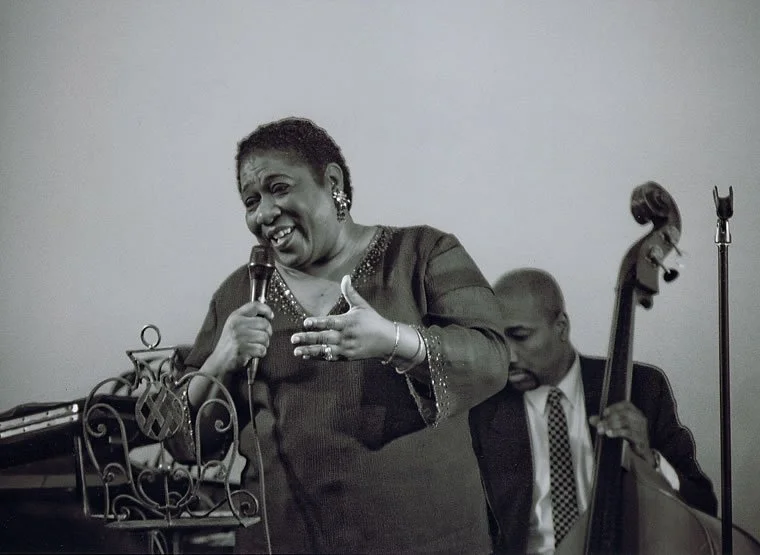 A woman singing and smiling into a microphone with a man playing a double bass in the background.