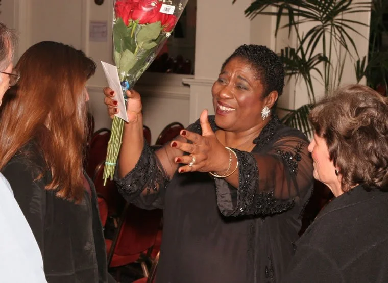 A woman giving a bouquet of flowers to another woman while three others look on, inside a room with plants and chairs.