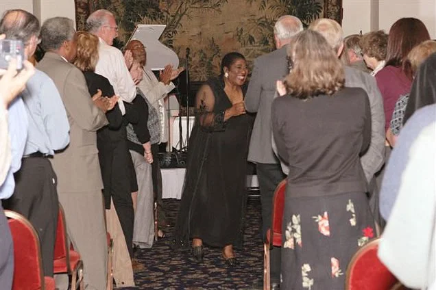 Group of people standing and clapping in a room, with a woman in black dress smiling at the center.