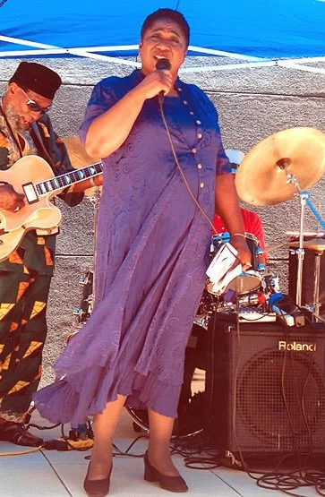 A woman singing into a microphone on an outdoor stage, with a man playing guitar beside her and musical equipment in the background.