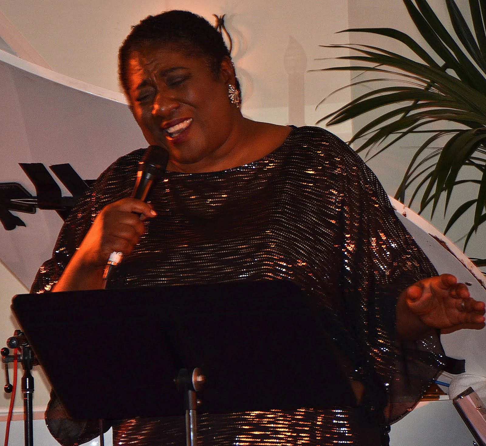 A woman singing into a microphone at an indoor event, wearing a black, shiny, sequined dress and earrings, with a green potted plant in the background.
