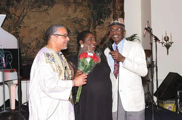 Three people smiling, one woman holding a bouquet of flowers, at a celebration or event in a decorated room.