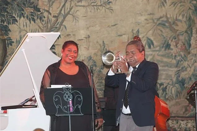 Jazz musicians performing live, with a woman singer smiling at a microphone, a man playing trumpet, and a white grand piano in the background, set against a decorative wall with a nature scene.
