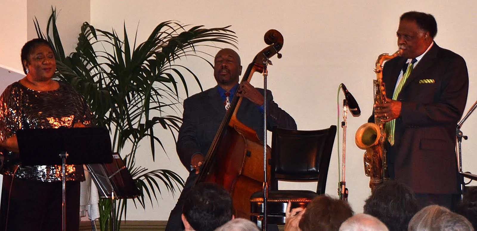 Jazz band performing on stage with three musicians: a woman singing, a man playing double bass, and a man playing saxophone, with audience members watching.
