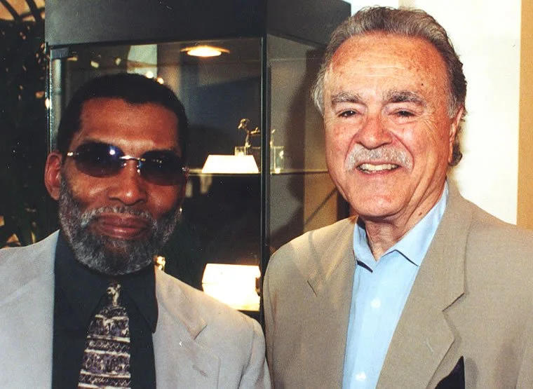 Two men in formal attire standing side by side indoors, one wearing a suit with a tie and sunglasses, the other in a suit without glasses, smiling.