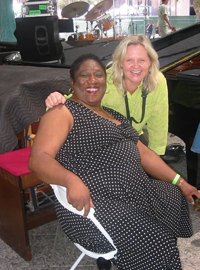 Two women smiling and posing together indoors, one sitting in a chair wearing a black polka dot dress, the other standing next to her wearing a light green jacket. There is a piano and musical instruments in the background.