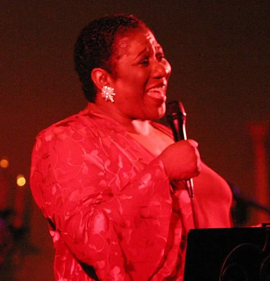 A woman singing into a microphone during a performance, wearing a red lace dress and large floral earrings.