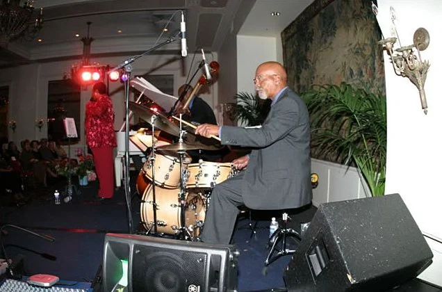 An elderly man playing drums during a live jazz performance at an indoor event.