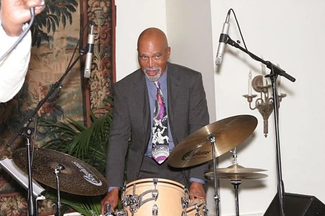 A man in a suit and tie playing drums with microphones set up in a room with a plant, tapestry, and wall sconce.