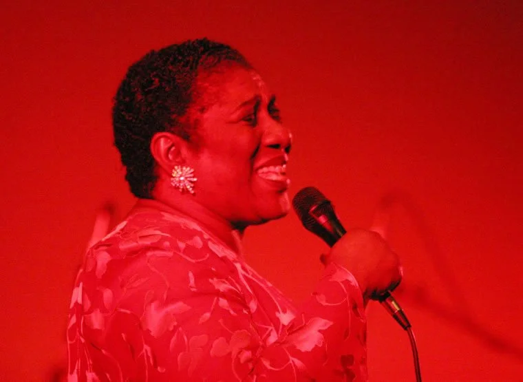 A woman with short curly hair singing into a microphone, wearing floral earrings and a floral dress, with a red background.