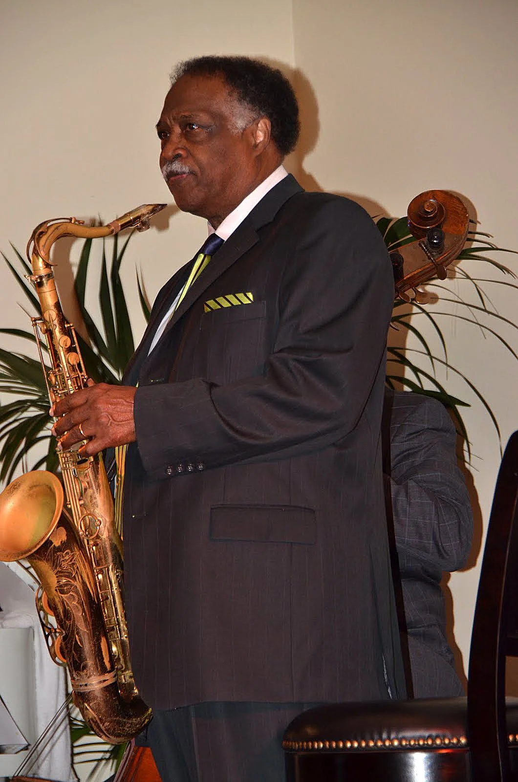 An African American man dressed in a black suit with yellow accents, playing a saxophone. He is indoors with a potted plant in the background.