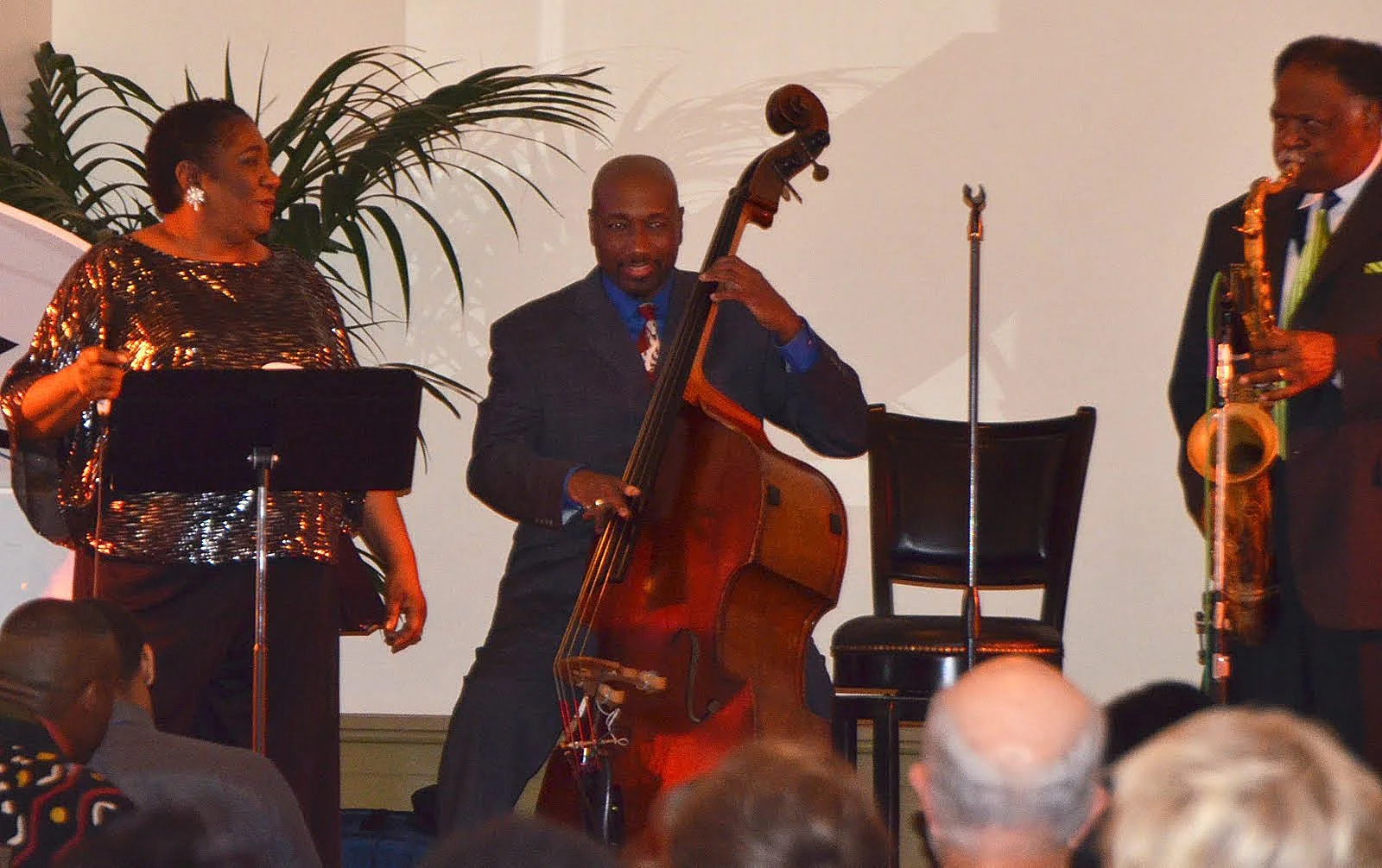 Jazz band performing at an event, with a female vocalist, a male double bassist, and a male saxophonist, all dressed in formal attire, in front of an audience.