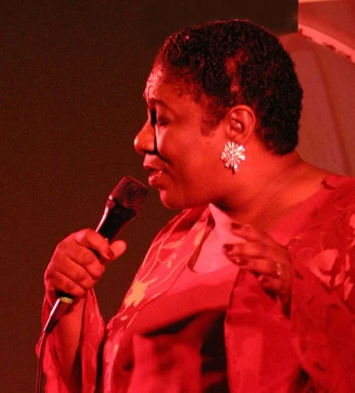 A woman with short curly hair and large earrings is holding a microphone and speaking on stage with a red backdrop.