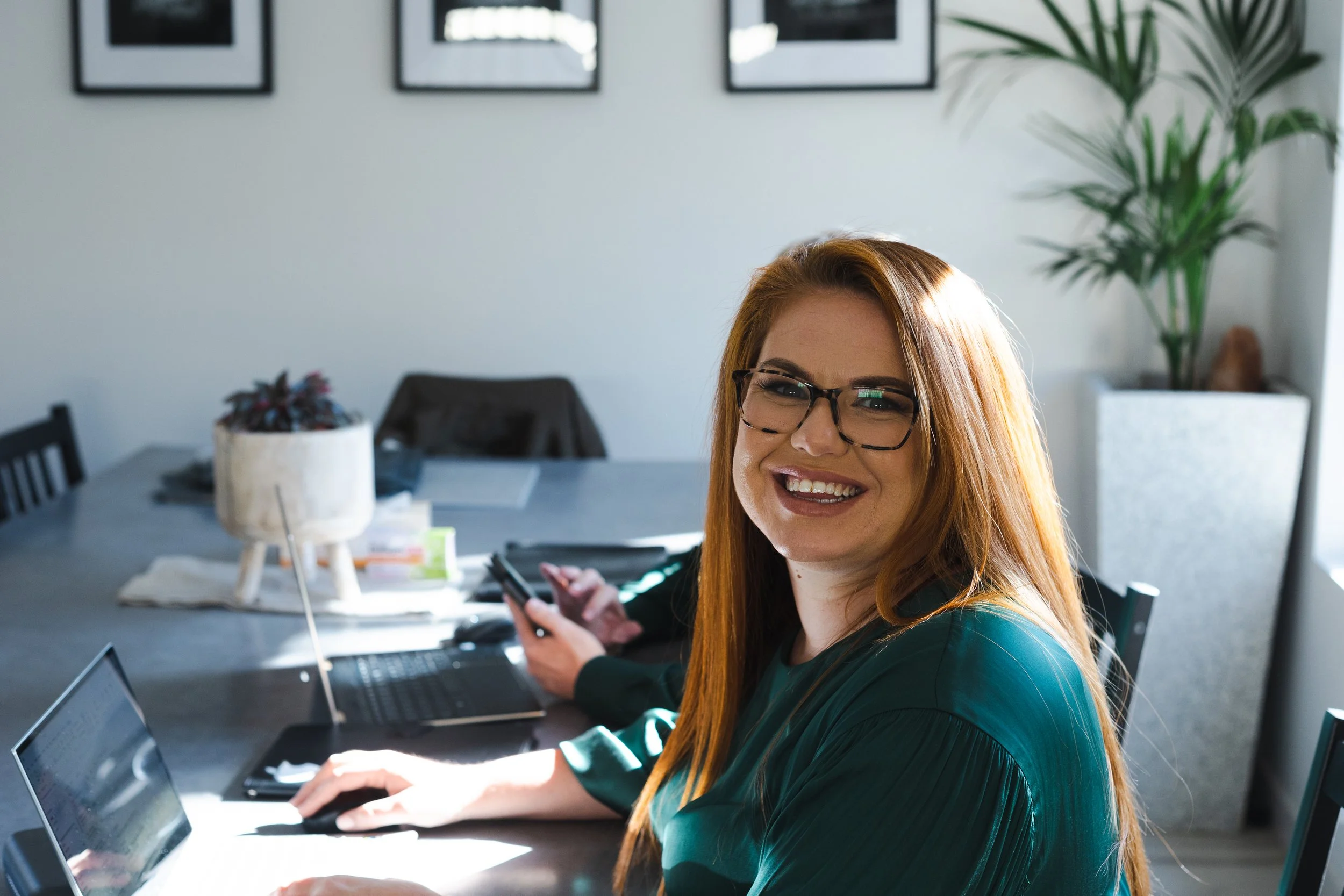 Woman with glasses smiling, sitting at a desk with a laptop and phone in a modern office, with framed pictures on the wall and a tall plant in the background.