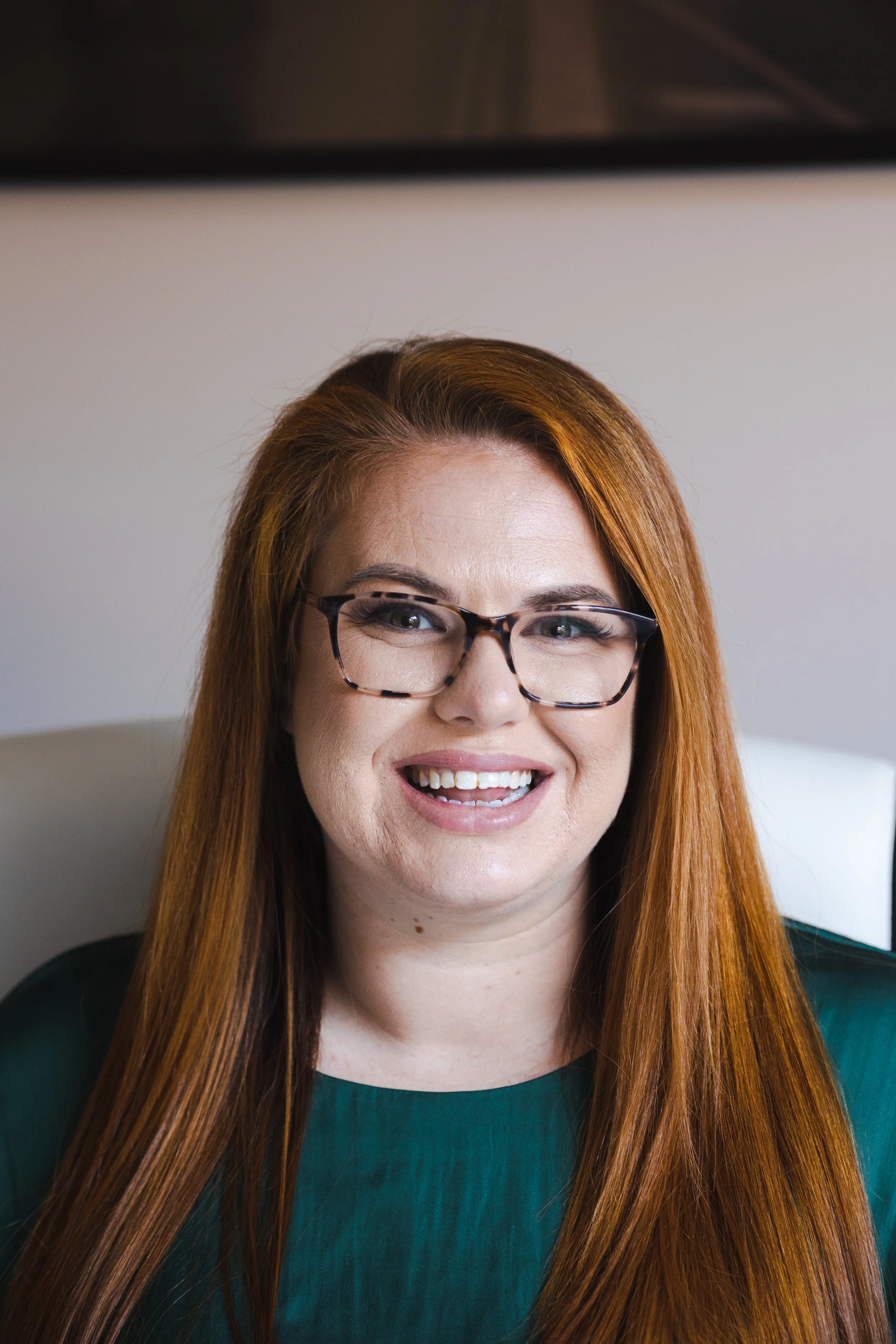 Close-up of a woman with red hair, wearing glasses, smiling, and wearing a dark green top, sitting in an indoor setting.