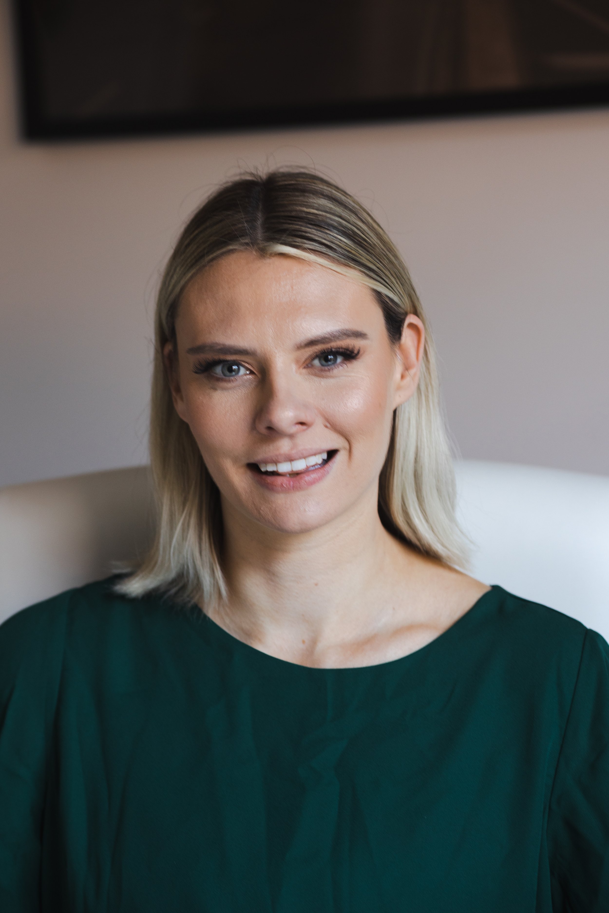 A woman with blonde hair and blue eyes smiling, wearing a dark green top, sitting in a room with a light-colored wall and a black-framed picture or window