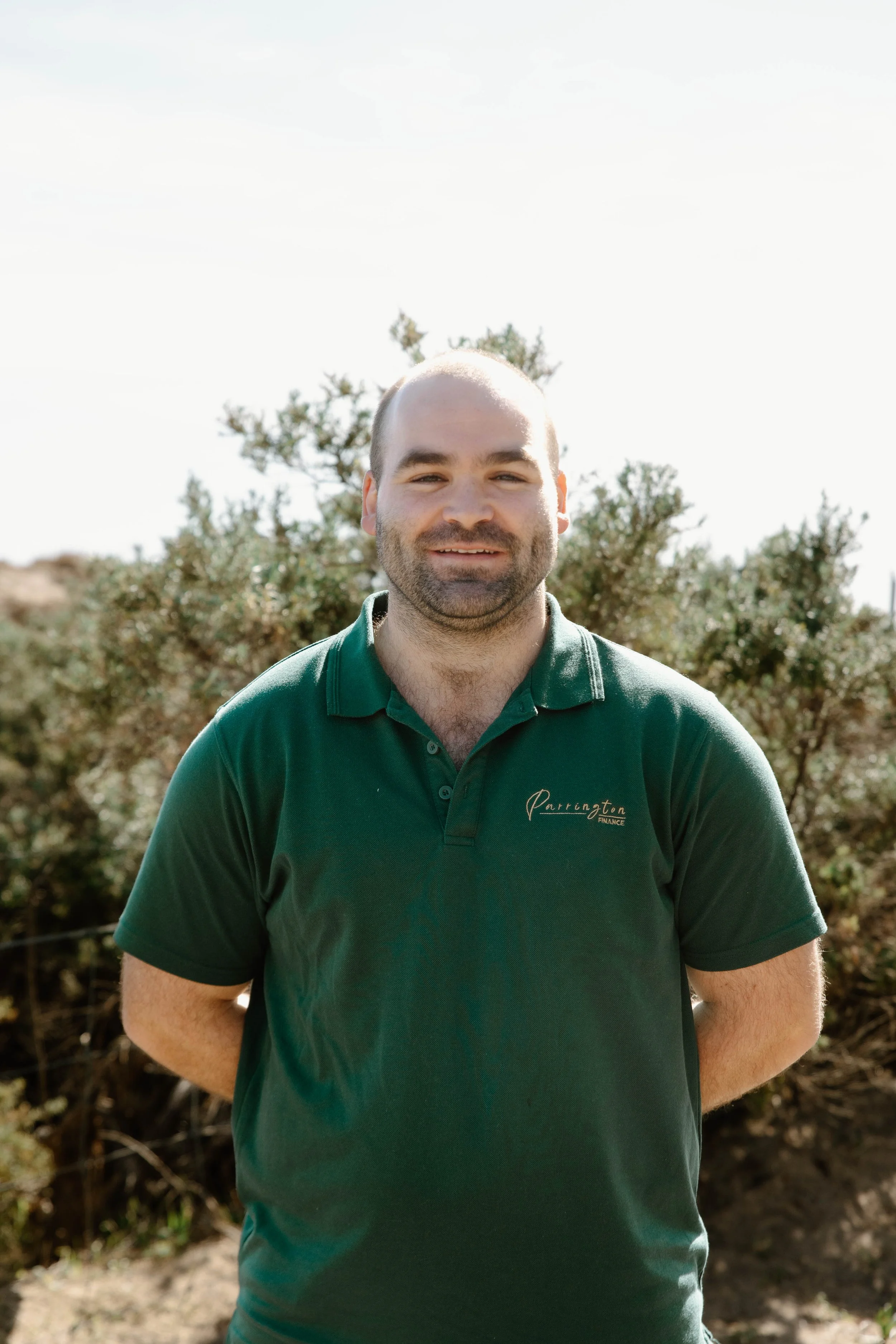 A man with a beard and short hair smiling outdoors, wearing a dark green polo shirt with the embroidered words "Partington Finance" on it, standing in front of bushes and trees with a bright sky overhead.