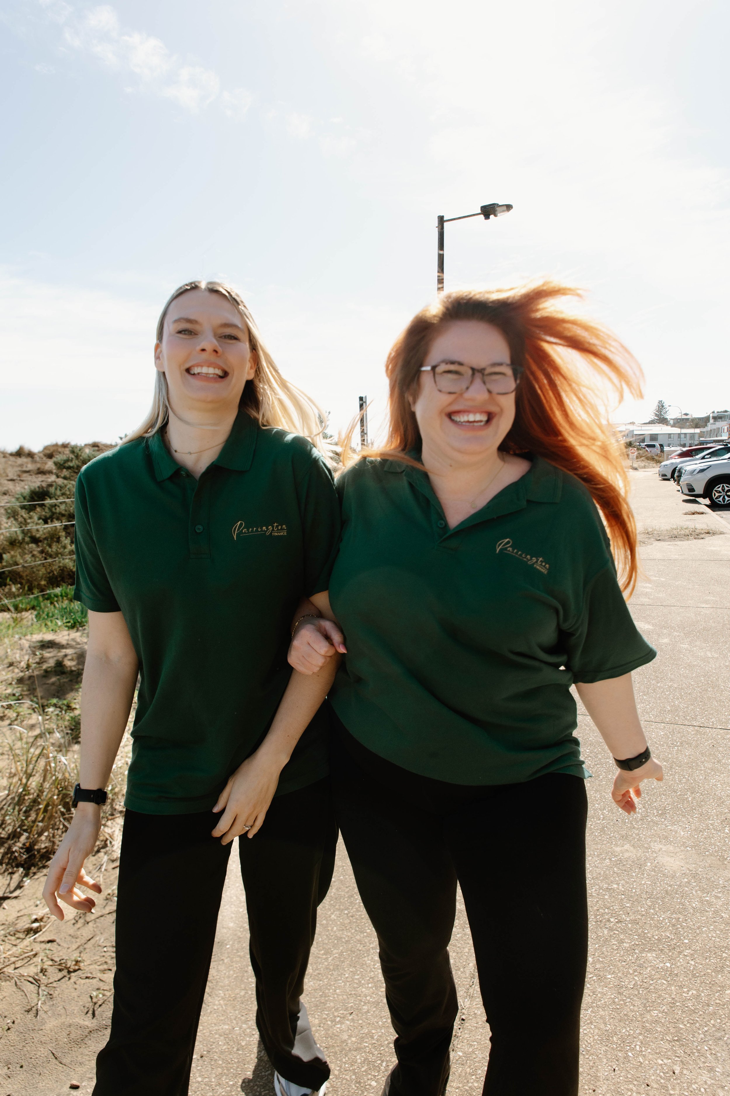 Two women smiling and walking outdoors on a sunny day, wearing matching dark green polo shirts, with one woman having red hair and glasses and the other with blonde hair, near a parking lot and street lamp.