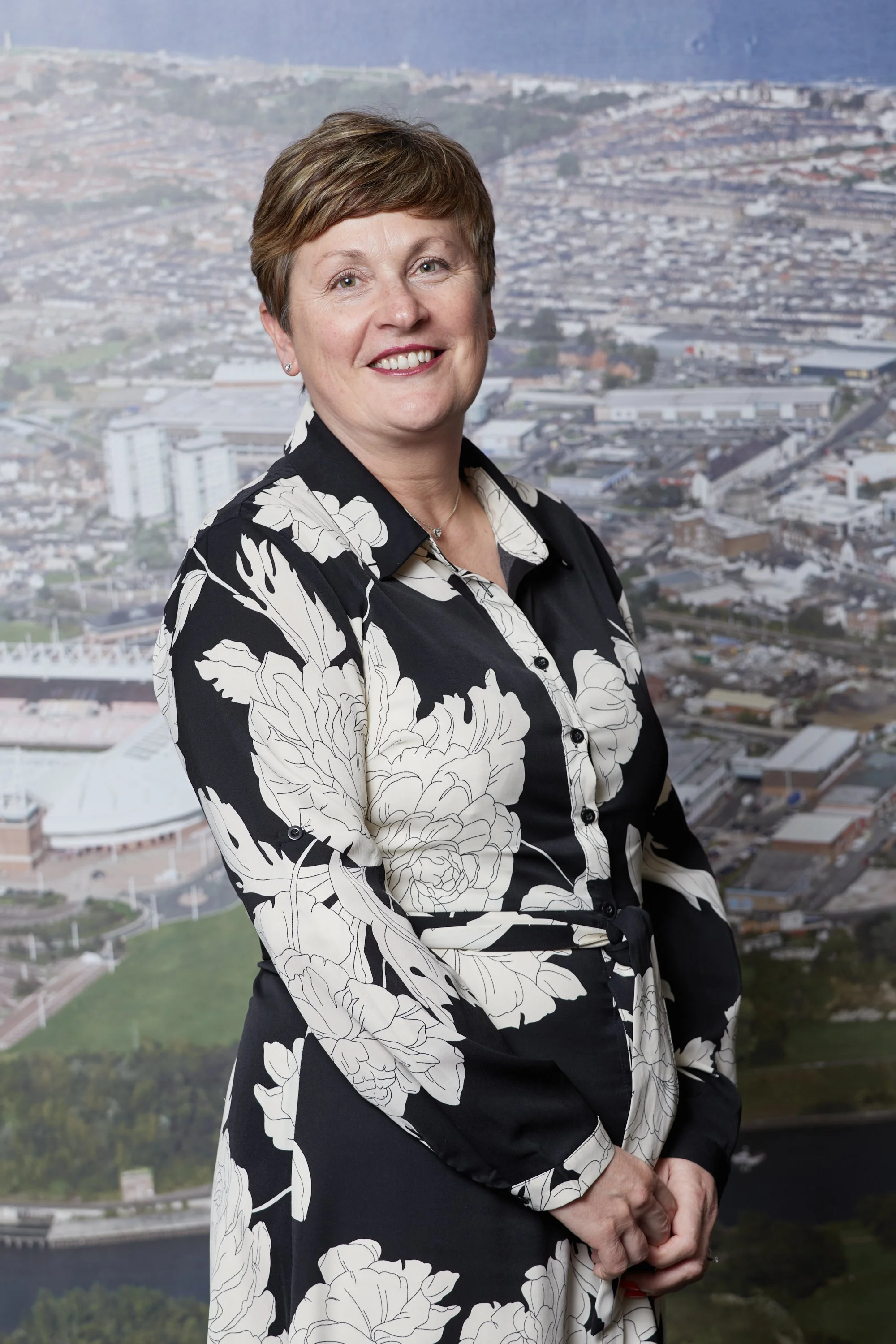 A woman with short brown hair and a black and white floral dress smiling in front of a cityscape view.