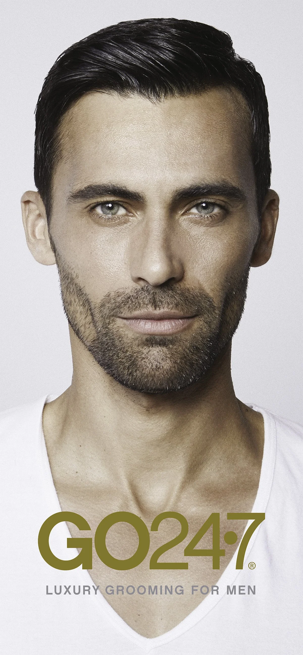 Close-up of a handsome man with dark hair and a beard, wearing a white shirt, with gold text that reads 'GO24.7' and smaller text 'Luxury Grooming for Men'