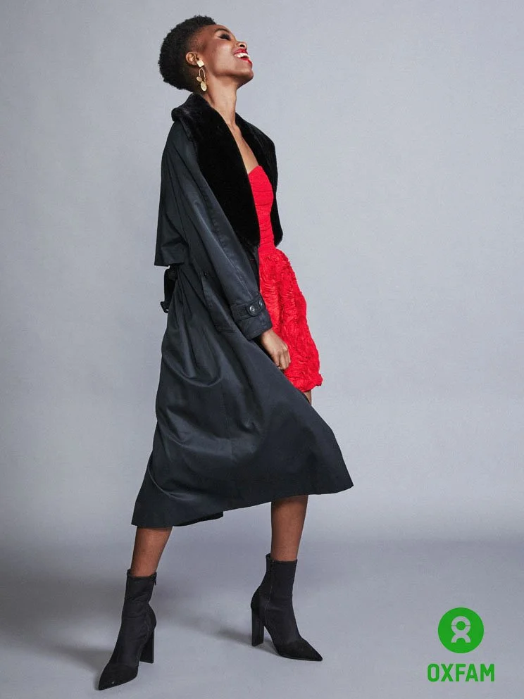 Woman in black coat, red dress, black high-heeled boots, earrings, smiling with head tilted back against a plain gray background, Oxfam logo.