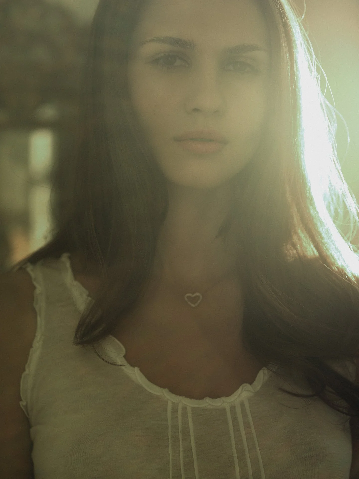 A woman with long dark hair, wearing a white top with ruffled straps and a heart-shaped necklace, stands outdoors in natural light.