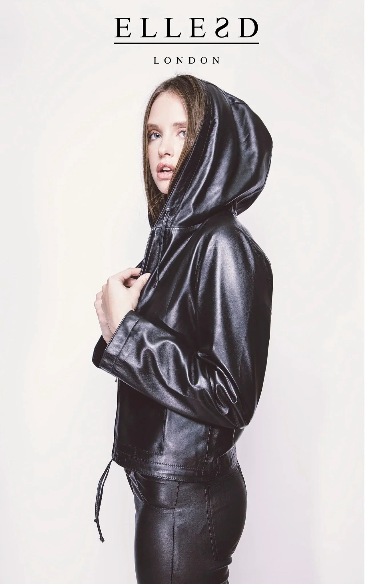 A woman wearing a black leather jacket with the hood up, standing against a plain light background.
