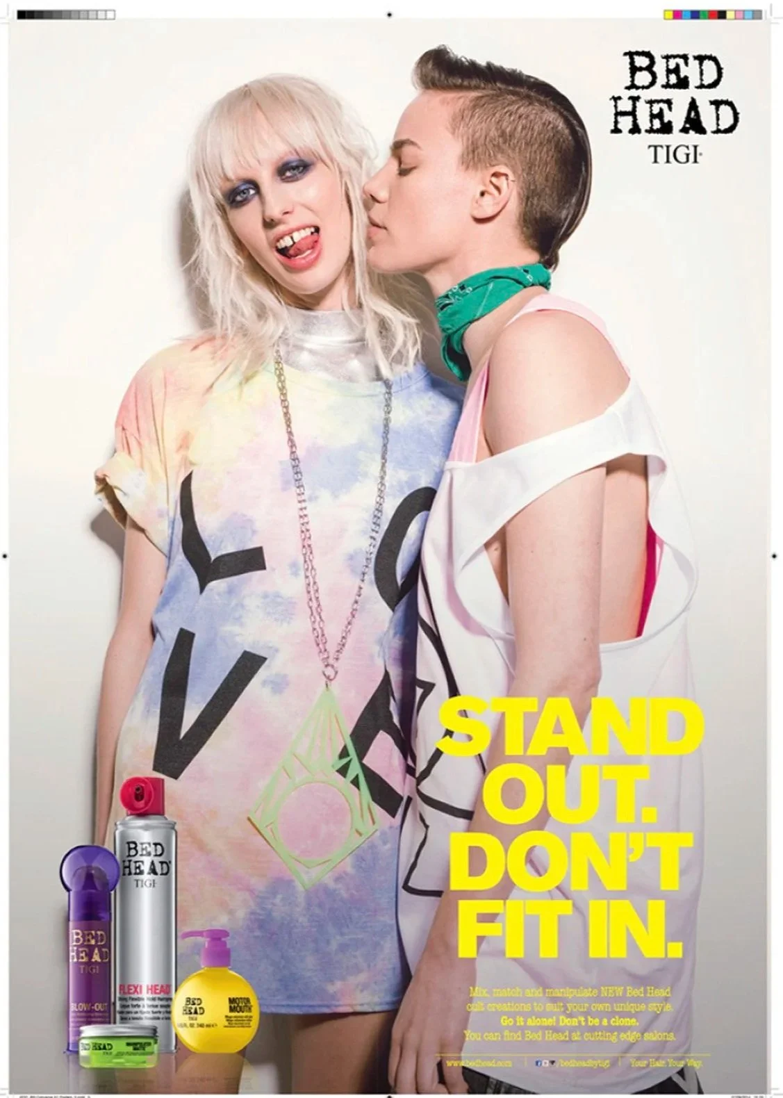 A promotional poster for Bed Head TIGI featuring two young women with alternative fashion styles. The woman on the left has platinum blonde hair, heavy eye makeup, and is sticking her tongue out. She wears a colorful tie-dye T-shirt with large black 