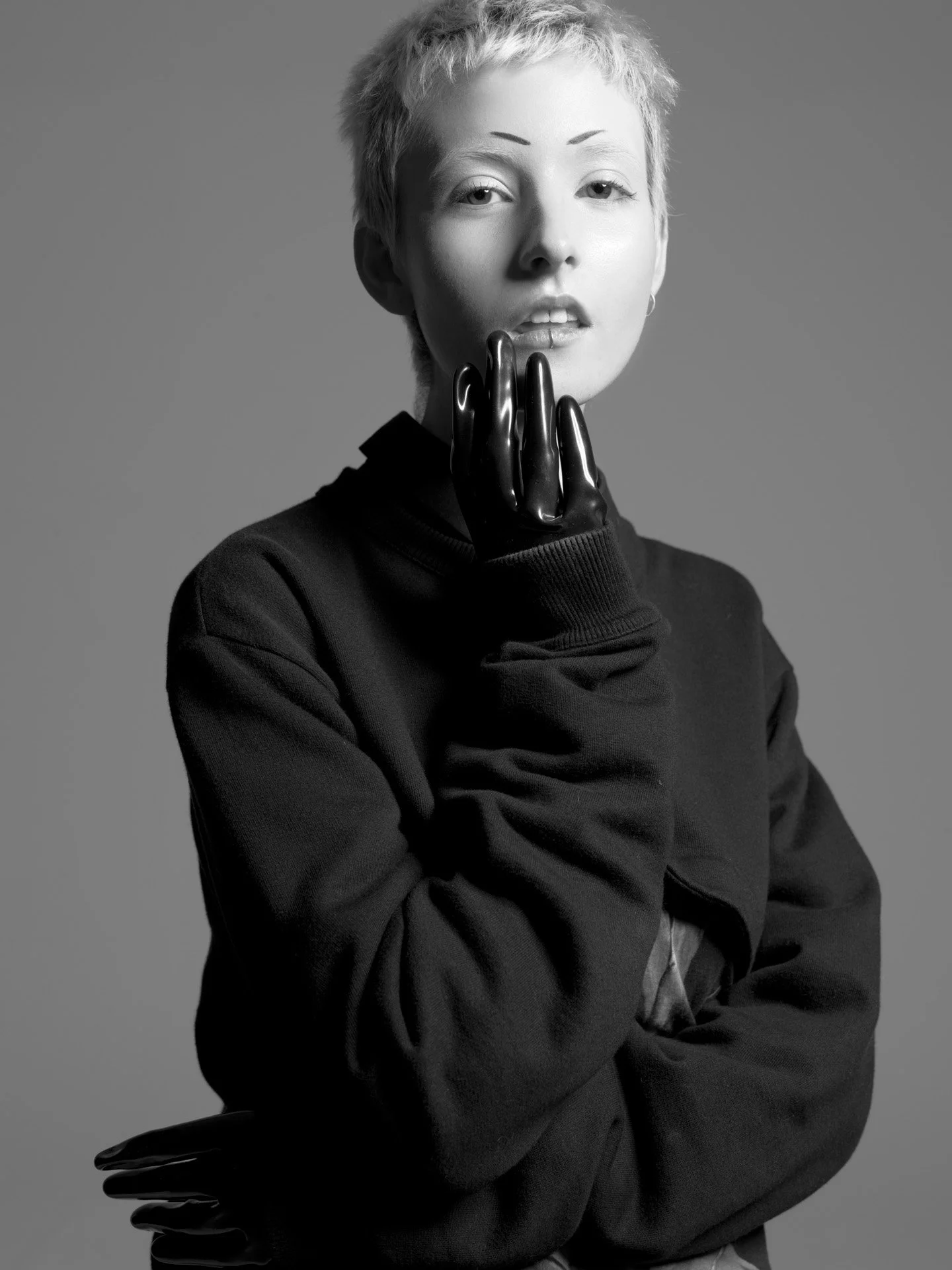 Black and white portrait of a young woman with short, spiky hair, wearing a dark sweatshirt, black gloves, and earrings, with her left hand resting against her face.