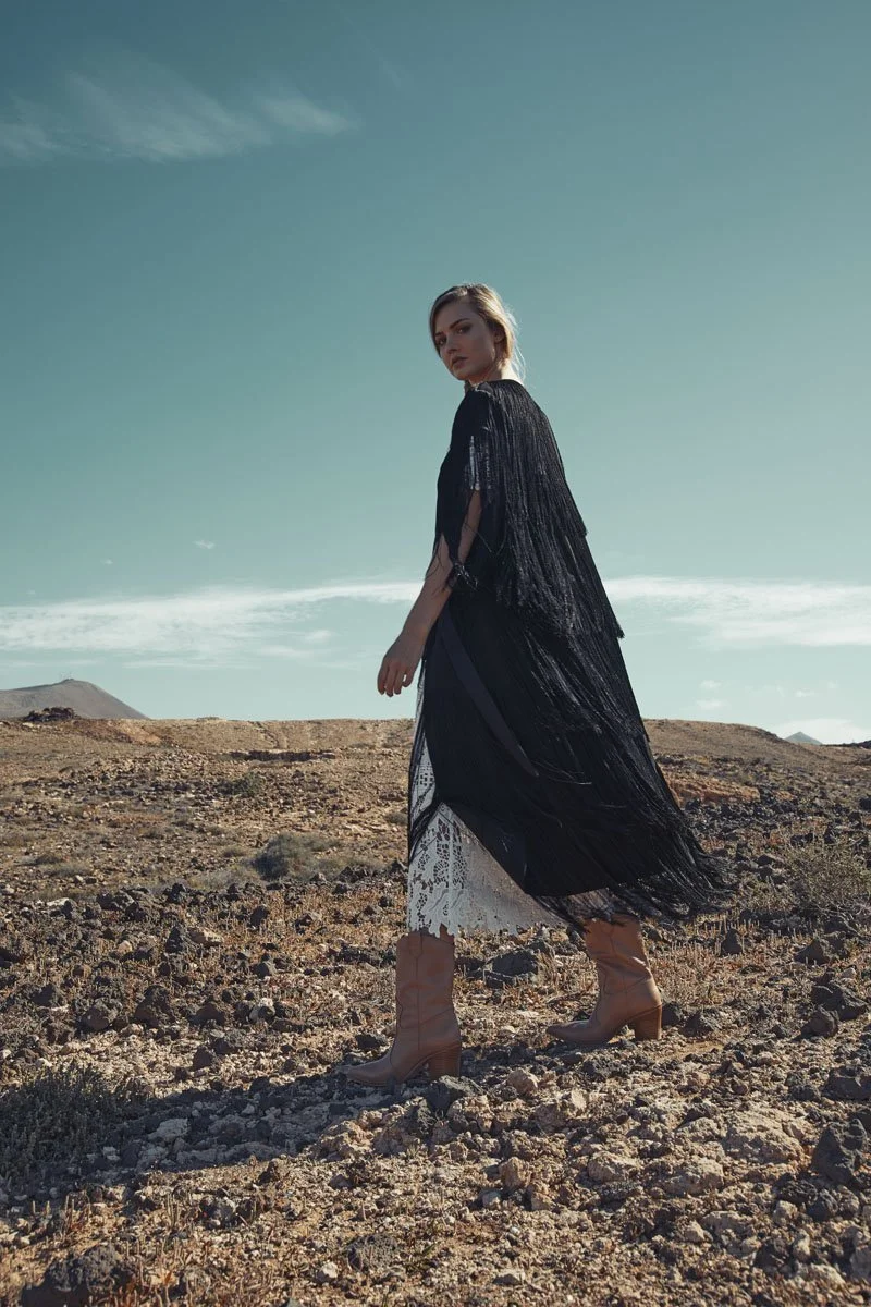A woman in a black and white dress with a long black fringe shawl standing in a barren, rocky landscape during daytime, with mountains and a partly cloudy sky in the background.