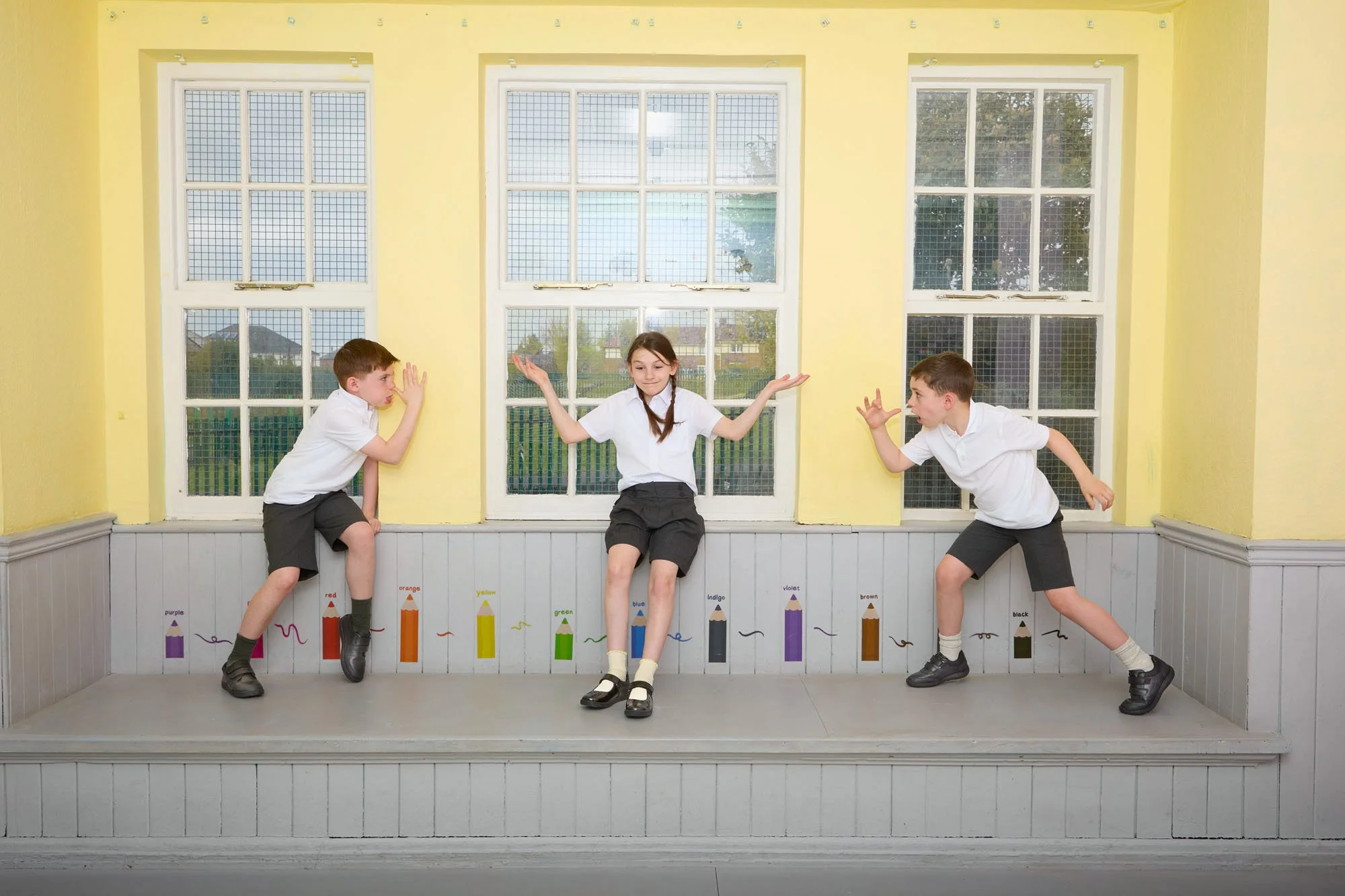 Three children dressed in school uniforms stand on a small stage in front of a yellow wall with a window. They are posing playfully, with the girl in the middle sitting on the stage, and the boys on either side leaning toward her. There are color-cod