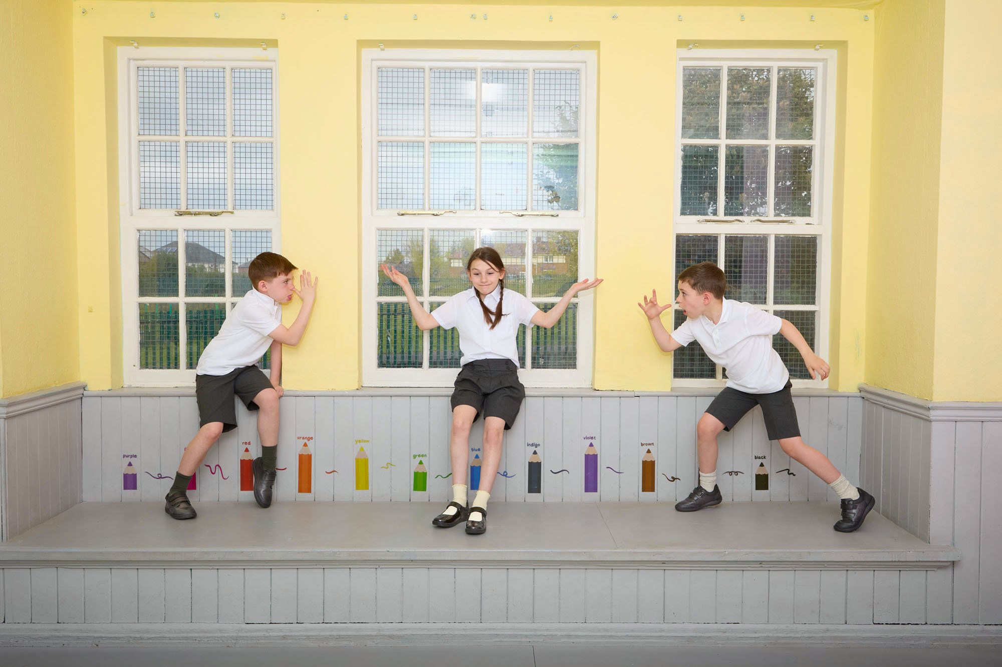 Three children dressed in school uniforms stand on a small stage in front of a yellow wall with a window. They are posing playfully, with the girl in the middle sitting on the stage, and the boys on either side leaning toward her. There are color-cod