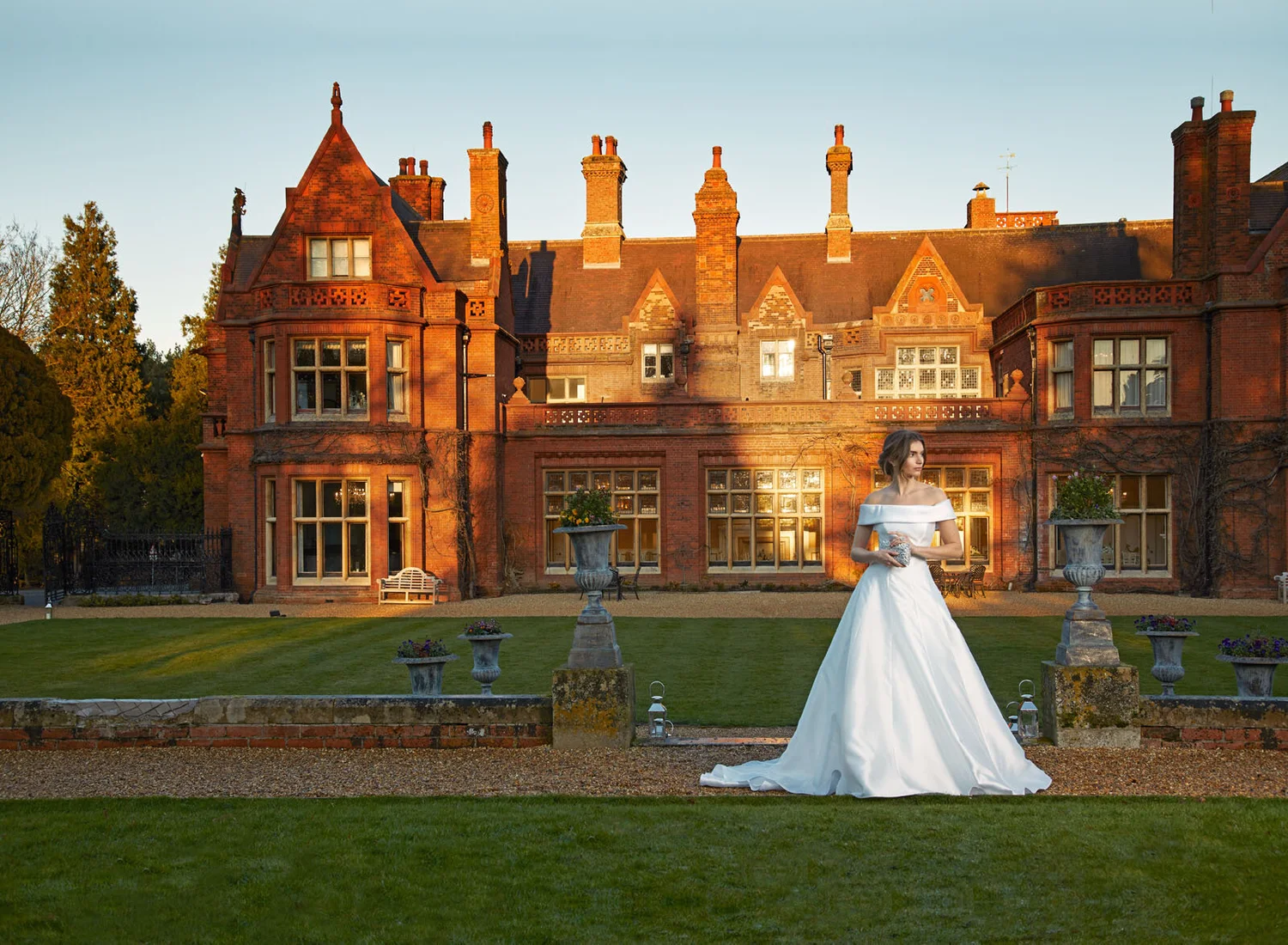 A woman in a white wedding dress standing on a lawn in front of a large, old brick mansion at sunset.
