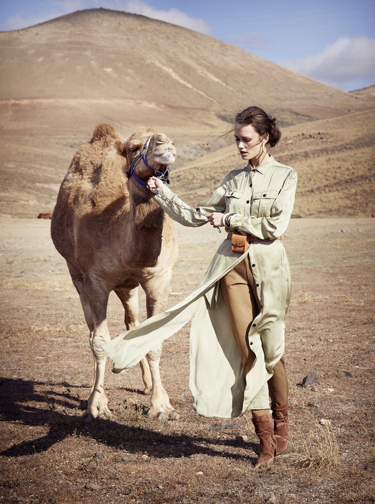 A woman holding a camel's reign in a desert landscape with rolling hills in the background.