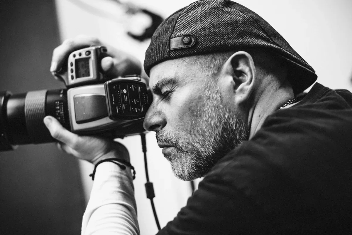 A man with a beard wearing a cap and a chain, with his eyes closed, holding a professional camera close to his face, appearing to be taking a photograph.