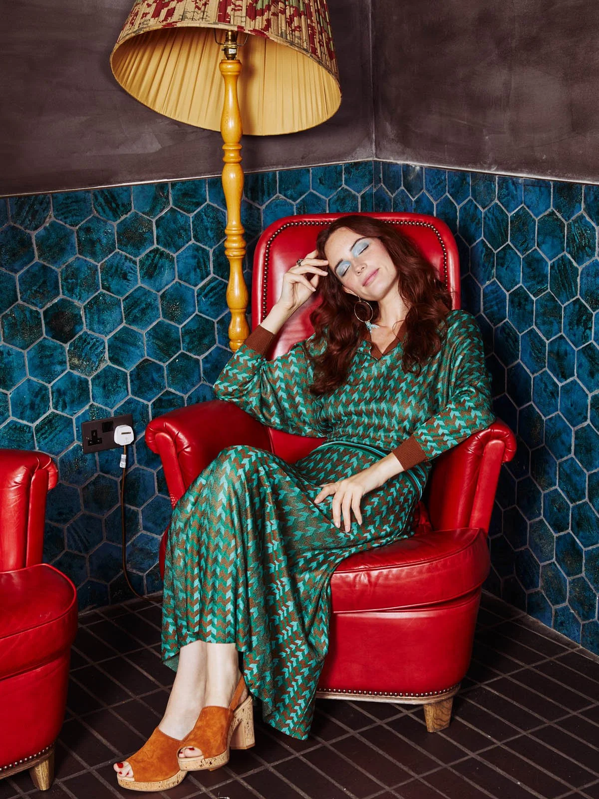 Woman with long red hair and closed eyes sitting in a red leather armchair with her hand on her forehead, wearing a teal and brown patterned dress and orange platform shoes, in a room with dark hexagonal tiled walls, a vintage floor lamp, and an elec