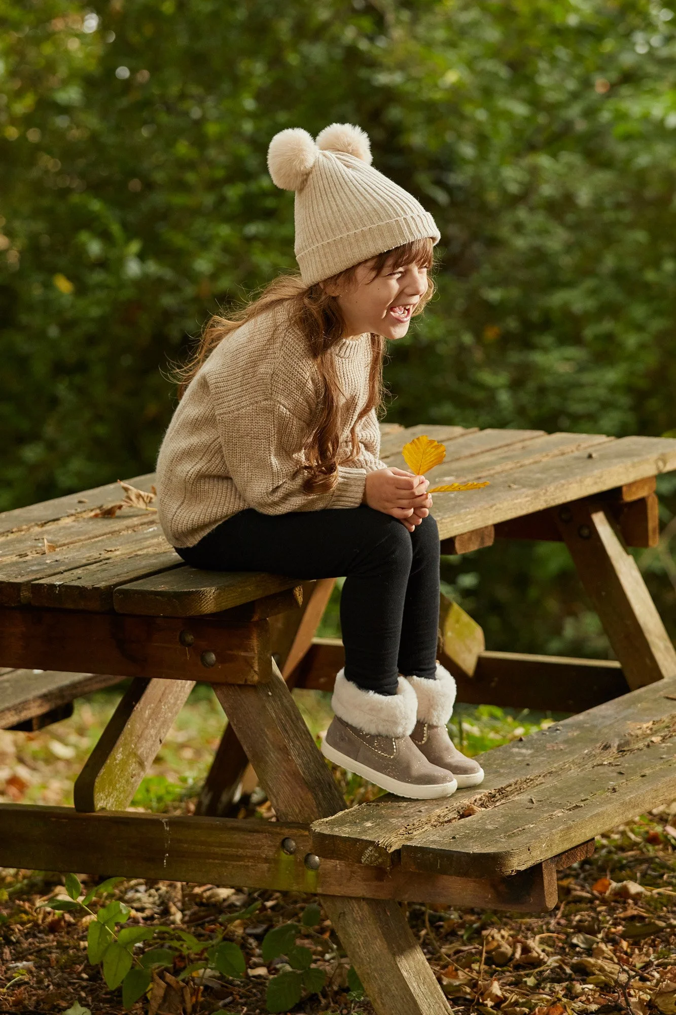 A young girl sitting on a wooden picnic table outdoors during fall, holding a yellow leaf, wearing a cream-colored knit hat with pom-poms, a beige sweater, and beige boots with fur lining.