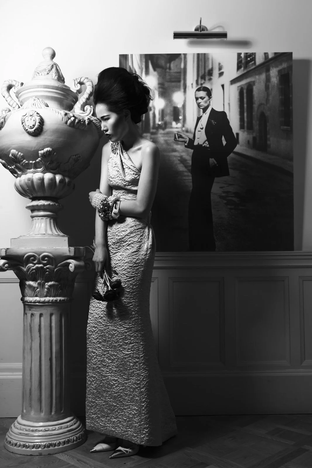 A black and white photo of a woman in a textured evening gown standing next to an ornate vase sculpture, holding a small bouquet. Behind her is a large photographic portrait of another woman in a tuxedo holding a glass, on a street scene backdrop.