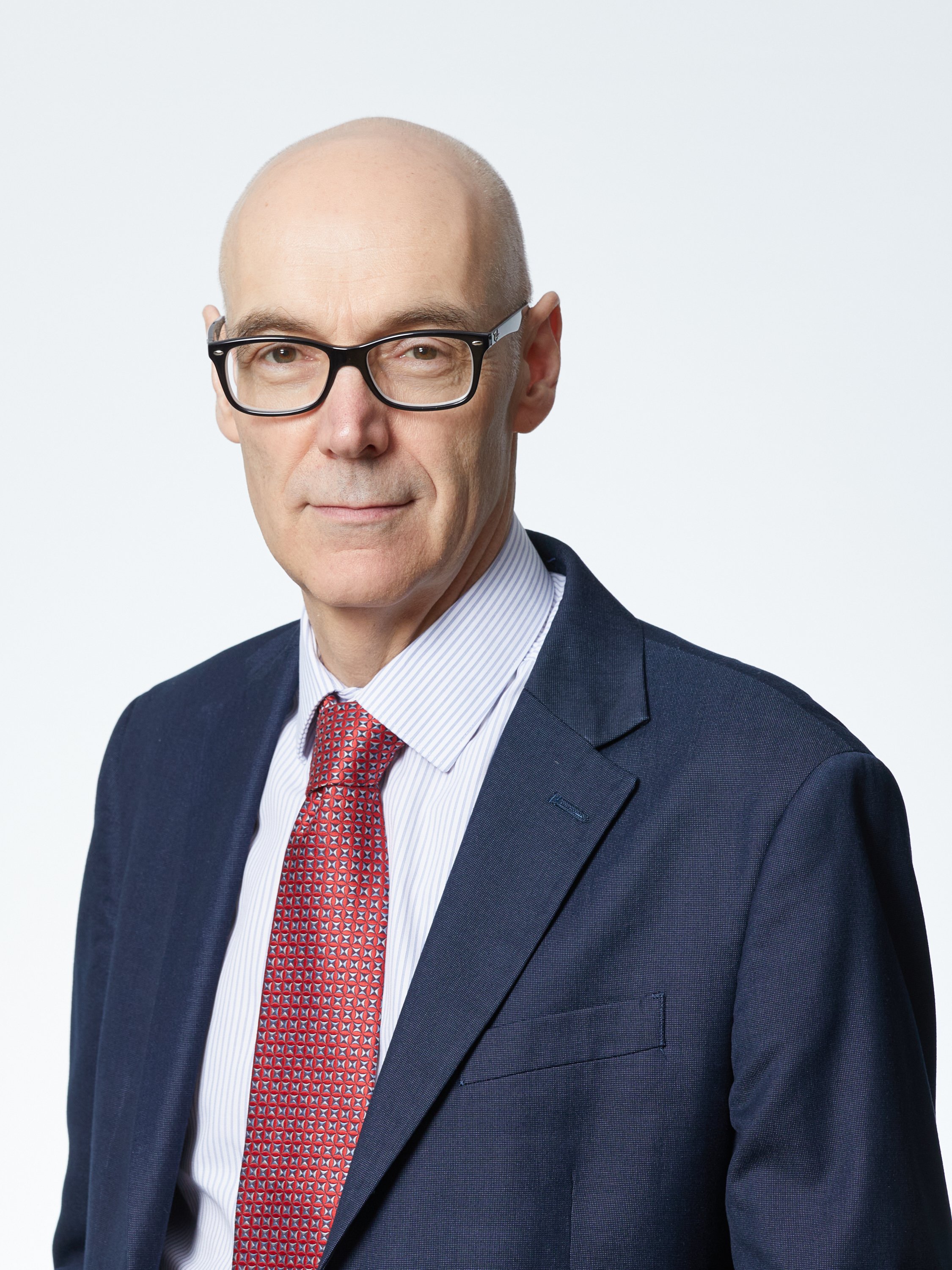 A professional man with glasses in a navy suit, white shirt, and red patterned tie poses against a plain white background.