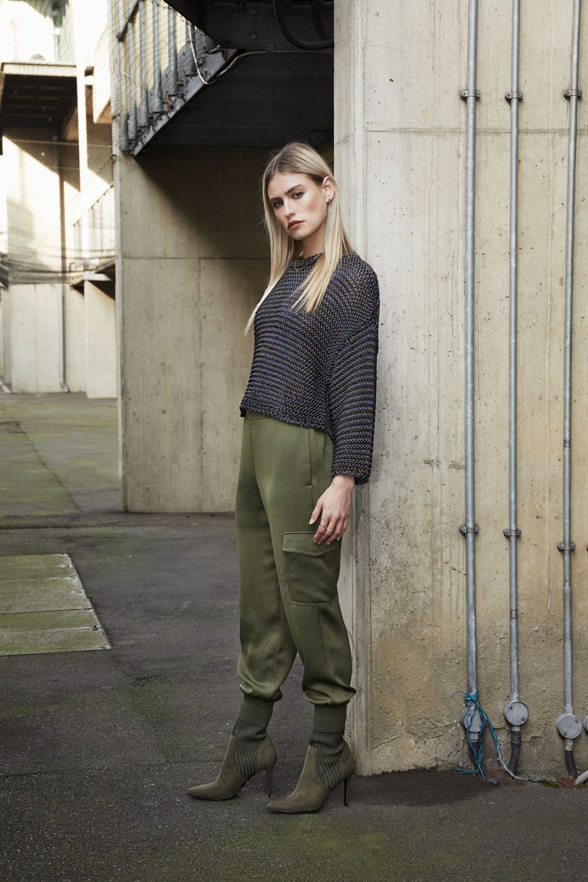 A young woman with blonde hair and dark lipstick leaning against a concrete wall in an urban setting, wearing a patterned black and gray sweater, olive green cargo pants, and high-heeled boots.