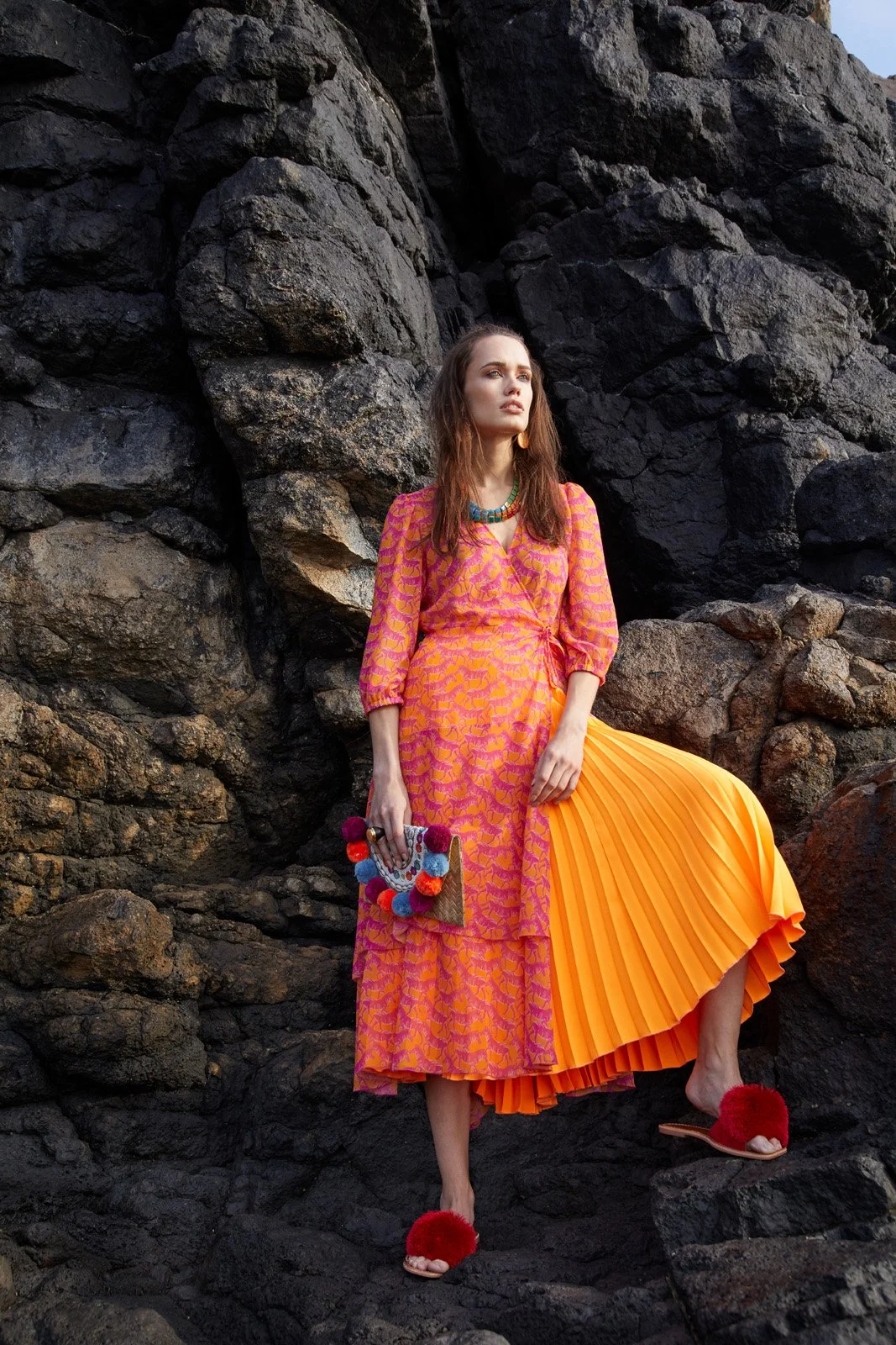 A woman standing against rocky terrain, wearing a bright pink and orange patterned dress with a pleated orange skirt, red fuzzy slippers, and holding a colorful beaded clutch.