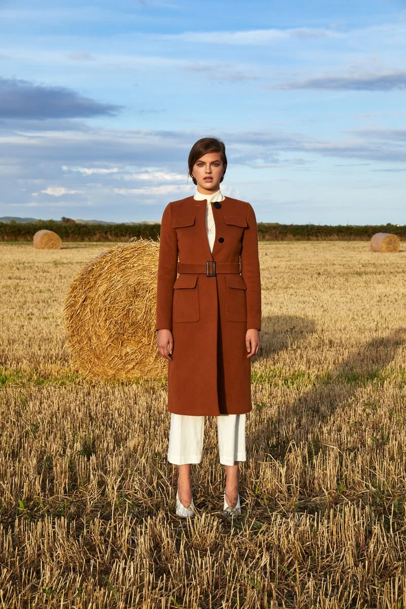 A woman stands in a wheat field with hay bales, wearing a long brown coat over a white dress, with a cloudy sky overhead.