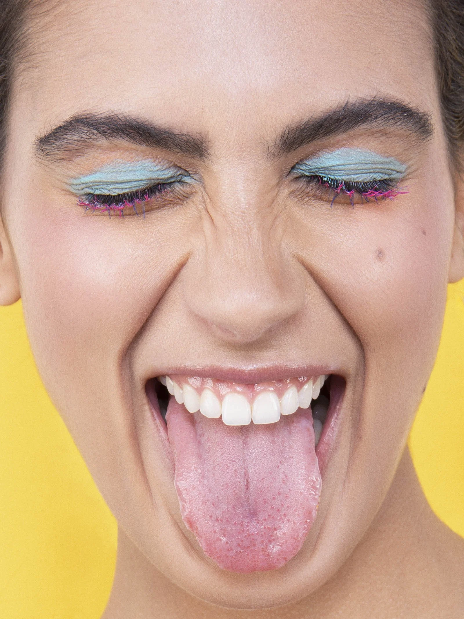 Close-up of a woman's face with blue eyeshadow, colorful eyelashes, and her tongue sticking out against a yellow background.