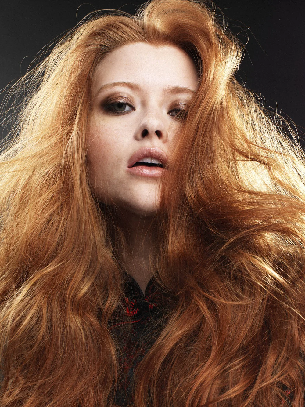 Close-up of a woman with long, voluminous red hair, wearing makeup with smoky eyes and slightly parted lips, against a dark background.