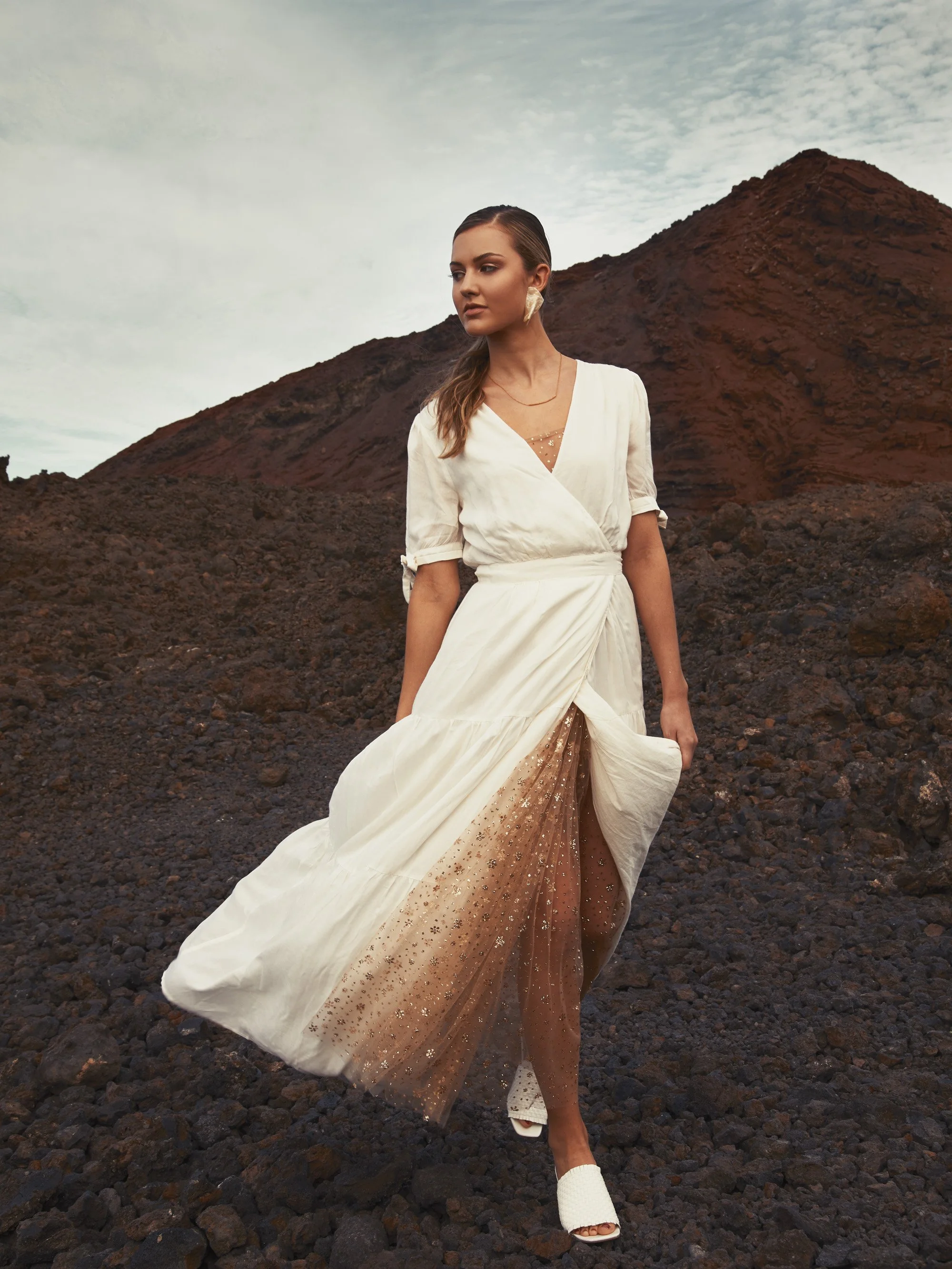 A woman in a white dress standing on rocky volcanic terrain with a reddish mountain and a cloudy sky in the background.
