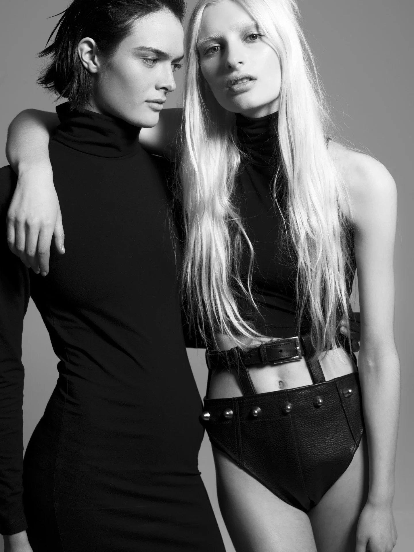Black and white photo of two young women standing close together, one with arm around the other's shoulder, both wearing dark clothing. The woman on the right has long, light hair and is wearing a black crop top and a black leather skirt with studs. 