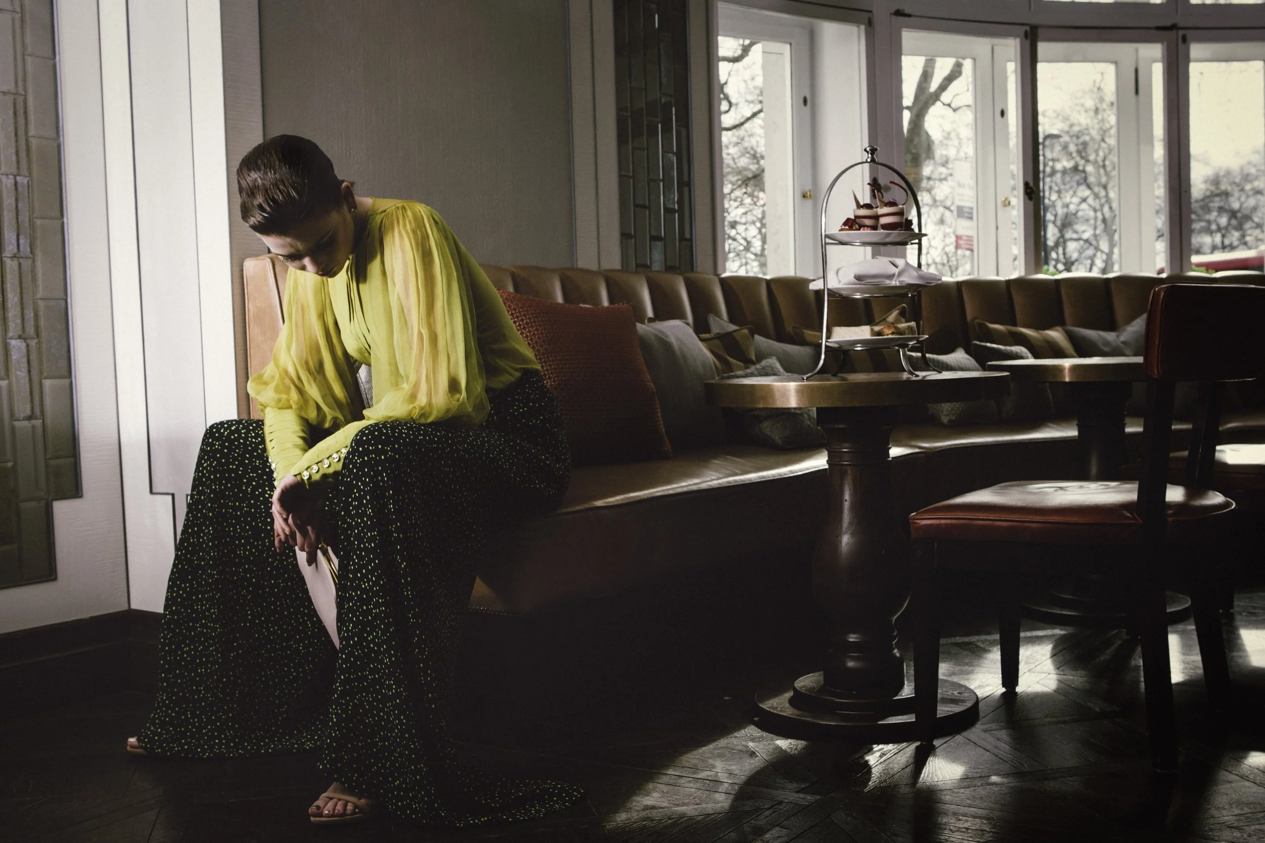 A woman dressed in a bright yellow blouse and black polka dot pants sitting in a restaurant or cafe, with a display of desserts and a window showing trees outside in the background.