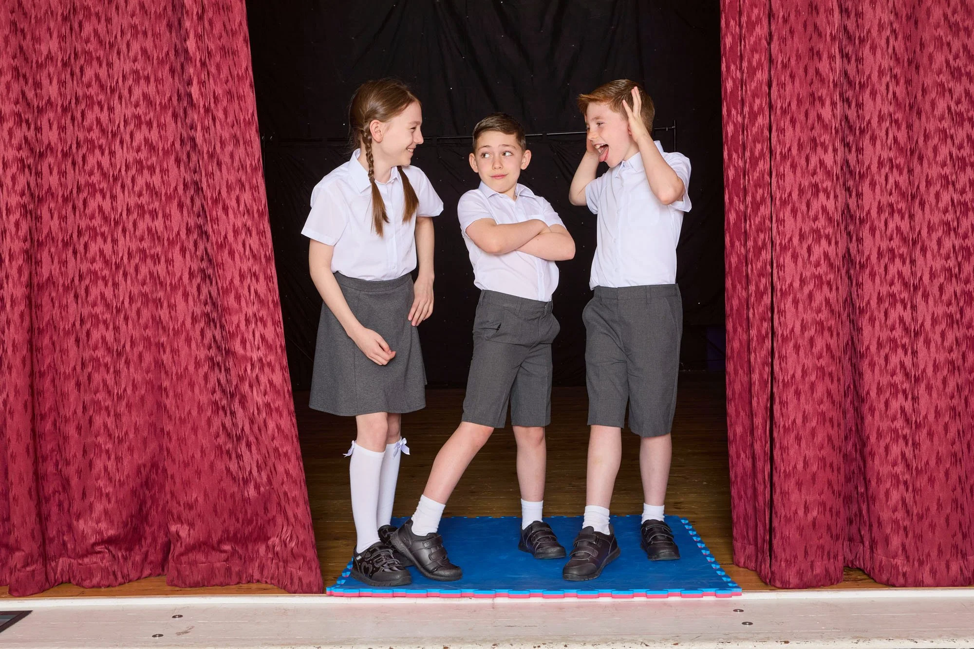 Three children dressed in school uniforms standing on a stage with red curtains on the sides. The children are engaging in a theatrical performance or school play, with the girl on the left smiling at the boy in the middle, who has his arms crossed a