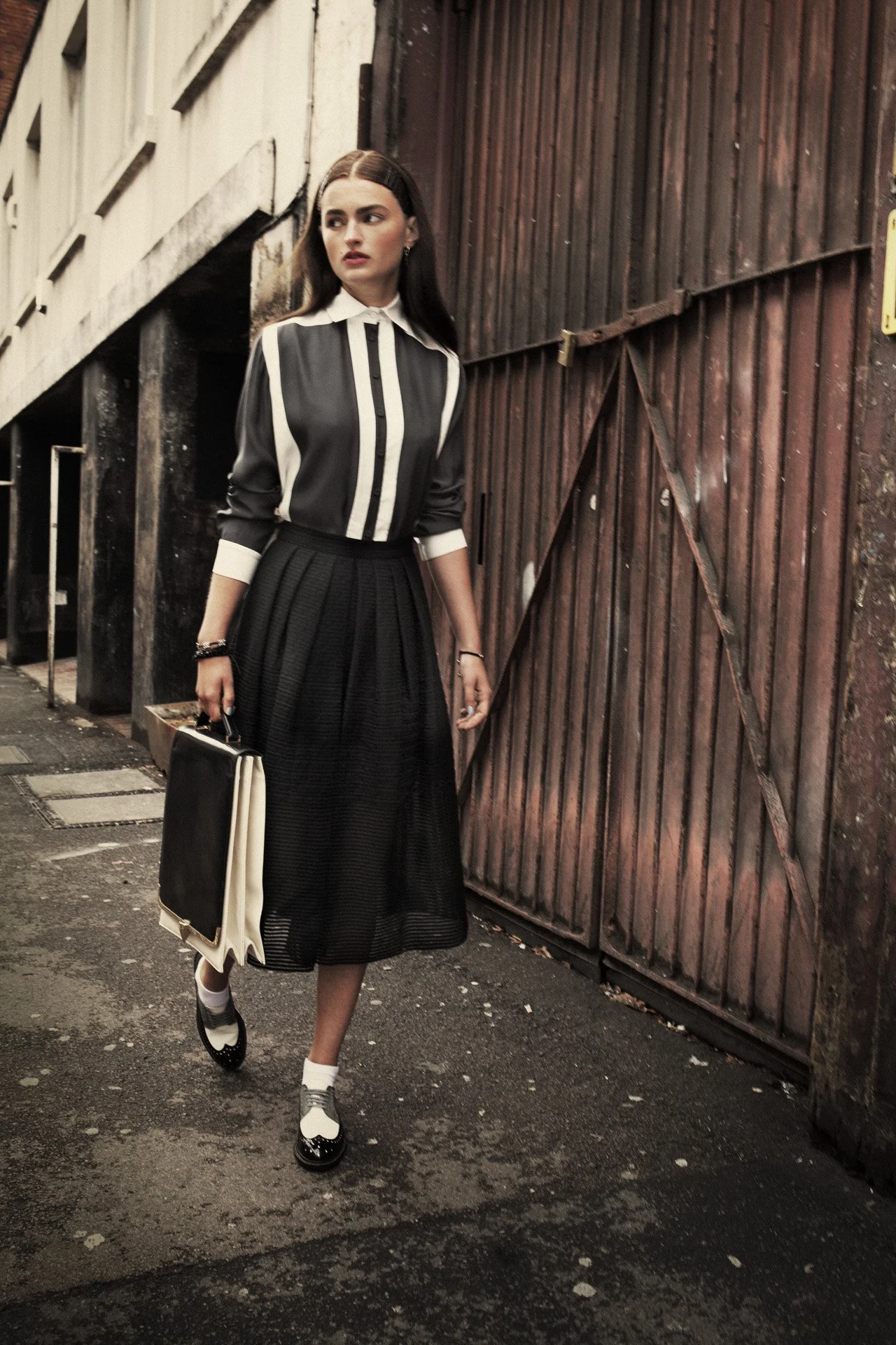 A young woman with long dark hair, wearing a black and white striped shirt, a black pleated skirt, and black and white shoes, holding a large black and white tote bag, walking along a city street with a wooden gate and building in the background.