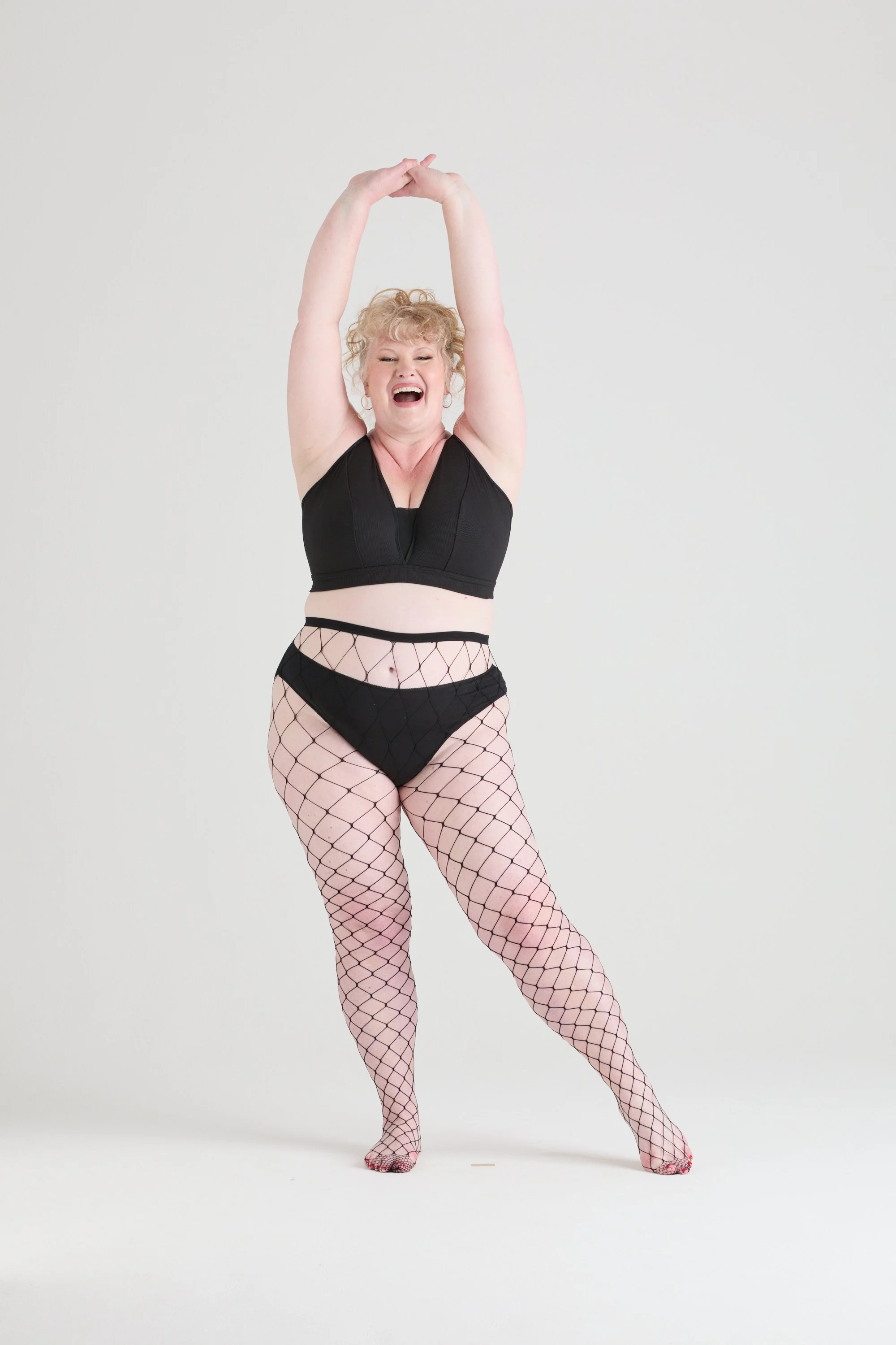 A woman with curly blonde hair wearing a black crop top, fishnet stockings, and lingerie, standing with her arms raised above her head against a plain, light-colored background.